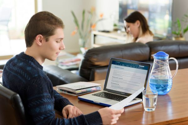 Tenant sitting at a table reading a letter with a laptop open to the SA Water website Information for Tenants page, a jug and glass of water on the table, and a girl sitting on the couch in the background.